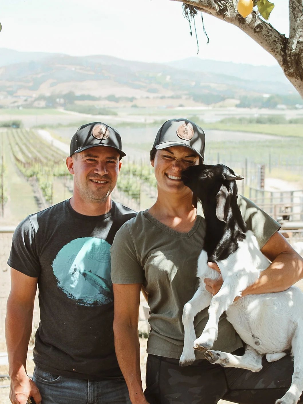Smiling couple holding a baby goat, vineyard behind them